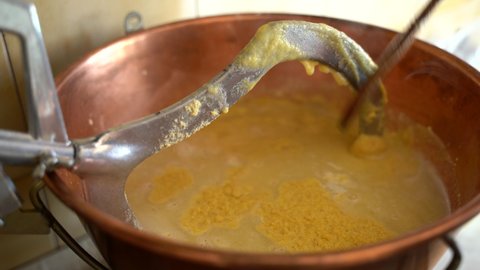 Man holding wooden spoon and stirring slowly yellow polenta porriage in copper cauldron on low fire at home. Preparation of traditional italian dish in northern part of country. Delicious winter