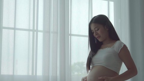 Young beautiful asian pregnant woman standing near window looking out the window at home