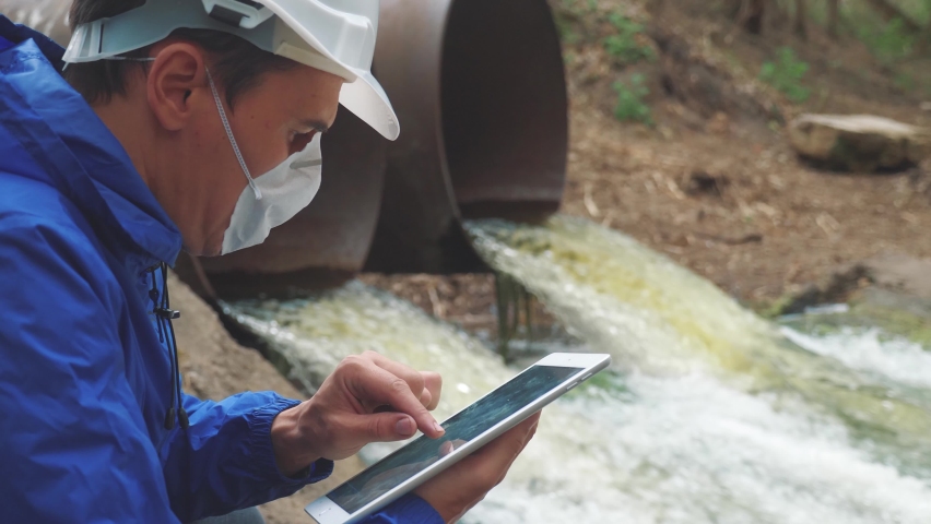 Environmental pollution. man chemist scientist in a respirator mask studies water pollution digital tablet and an white helmet stands next to the waste pipes. radiation waste concept. conservation | Shutterstock HD Video #1065187174