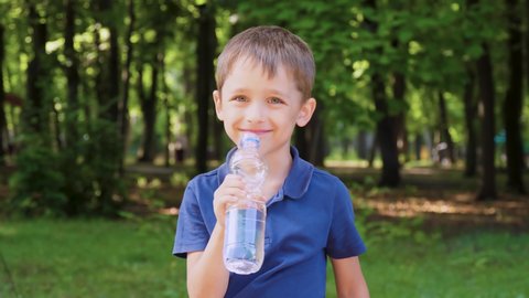 A happy child drinks water from a bottle on a sunny hot day in a park, against the background of a green forest. The concept of clean water, healthy lifestyle and environmental protection. A portrait