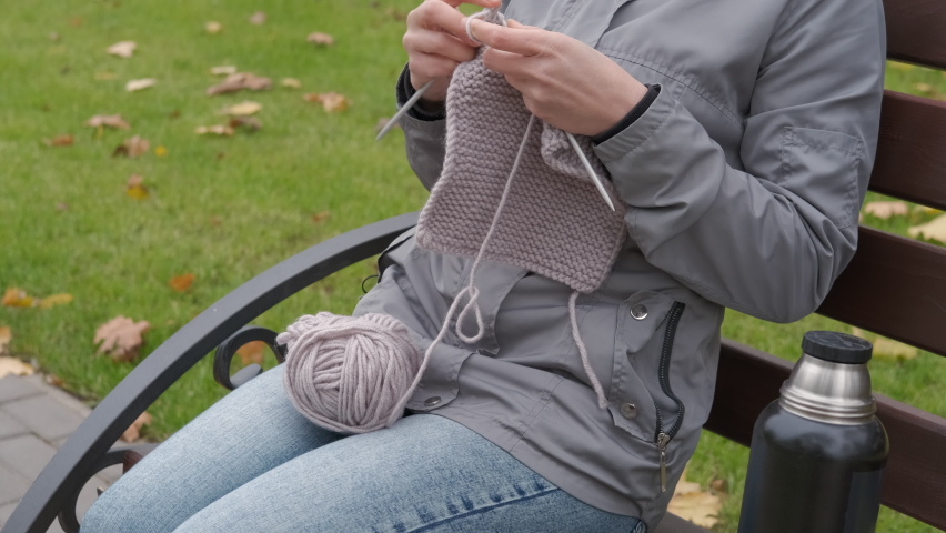 Sits with yarn in the park. A woman sits on the bench in the cold day and knitting by the thermos with tea.