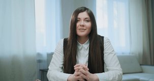 Young brunette fixes long loose hair and talks to corporate manager sitting on white sofa at home at online video conference