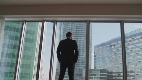 Silhouette of successful young businessman in suit is looking at window. He admires panoramic city view from his modern apartment, back view. A successful businessman looks at skyscrapers from his win