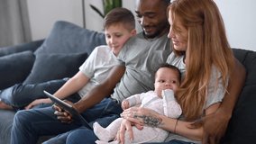 A multiracial family of four relaxing with a digital tablet sitting on the comfortable sofa, watching video, playing game on the touchscreen, laugh and have fun together