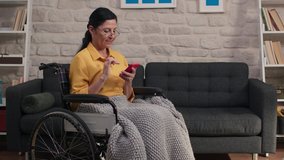 Old woman with glasses sitting in a wheelchair at home, reading the news on her mobile phone.Slow motion video.