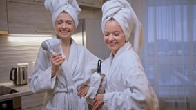 Two girl friends in white bathrobe dance, sing and dry hair with hairdryer, laugh. Young women have fun time. Funny video