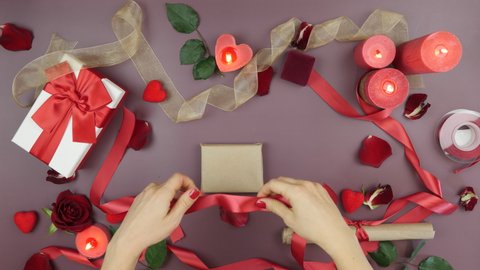 Female hands are tying red ribbon on gift box for St. Valentine's Day on festive table background with roses, candles, gifts and hearths. Woman is decorating gift box for present for Valentines Day