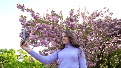 European woman move long hair by hand and take selfie, she stand against flowering Japanese cherry tree at Shinjuku Gyo-en park. Tourist lady enjoy beautiful spring time at Tokyo city