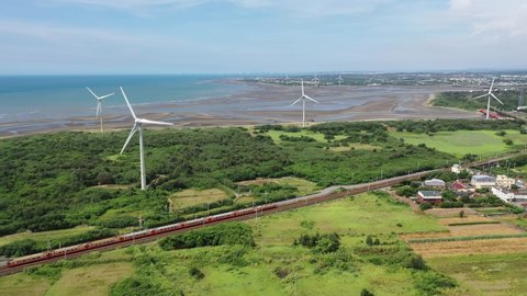 Aerial view of a Chu-Kuang Express train traveling thru green fields by a beach and giant wind turbines turning under blue sunny sky in a seaside park along the windy coast in Houlung, Miaoli, Taiwan