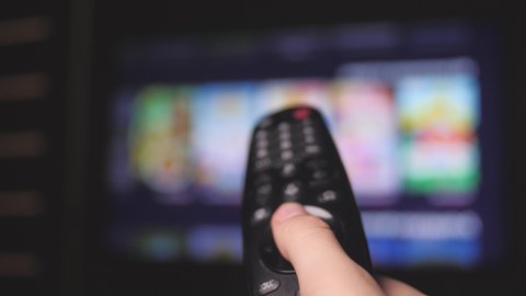 Man's hand selects internet tv channels with remote control, close-up. Person controls TV using a modern remote control. A man watches smart TV and uses black remote control. Blurry tv scrolls pages.