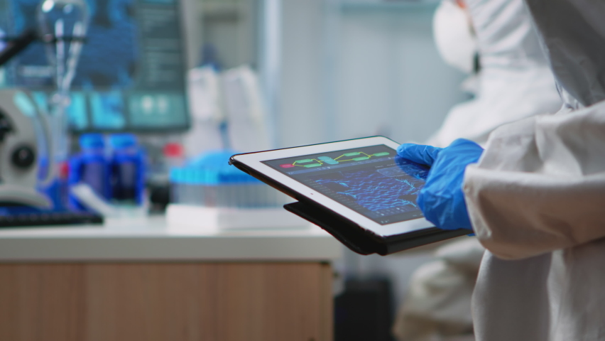 Back view of scientist in coverall analyzing virus evolution looking on digital tablet. Team of microbiologists conducting vaccine development using high tech for researching treatment against covid19 Royalty-Free Stock Footage #1065221062