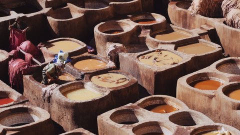 FEZ, MOROCCO - OCTOBER 2020: Elevated Handheld Panning Wide Shot Of Men Standing In The Dye Pots, Cutting And Preparing Leather In The Chouara Tannery, Fez, Morocco