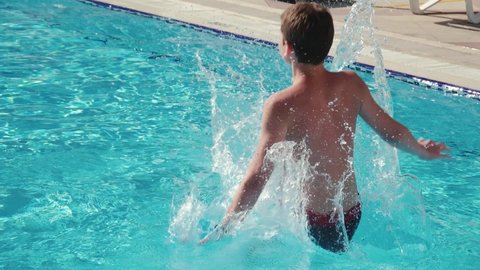 Unrecognizable guy jumping in pool and splashing crystal clear water in basin at sunny day. Boy relaxing during summer vacation, slow motion