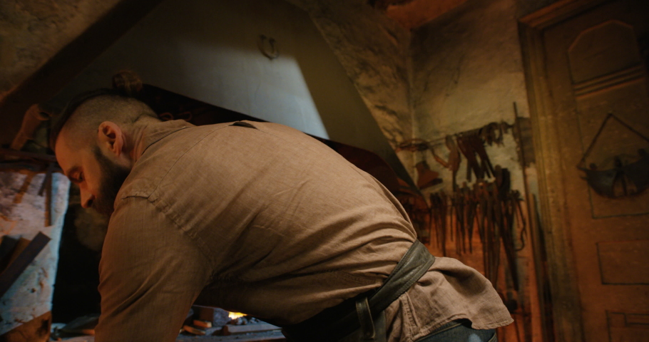 Young handsome male smith with long beard taking out molten glowing metal horseshoe from fire flame and looking at it with interest in antique smithy. Brutal forger working in workshop manufactory.