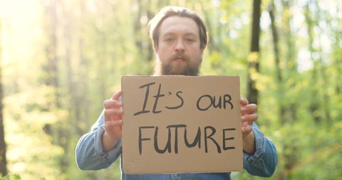Portrait shot of Caucasian young male eco acivist standing in green forest and holding poster with words It's our Future. Handsome man protesting for clean and safe environment.