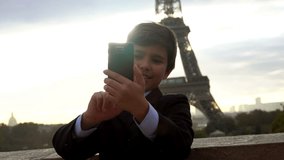 A cheerful teenage boy in a black suit is taking a photo on the phone on the background of the Eiffel tower at the sunrise, Paris, France
