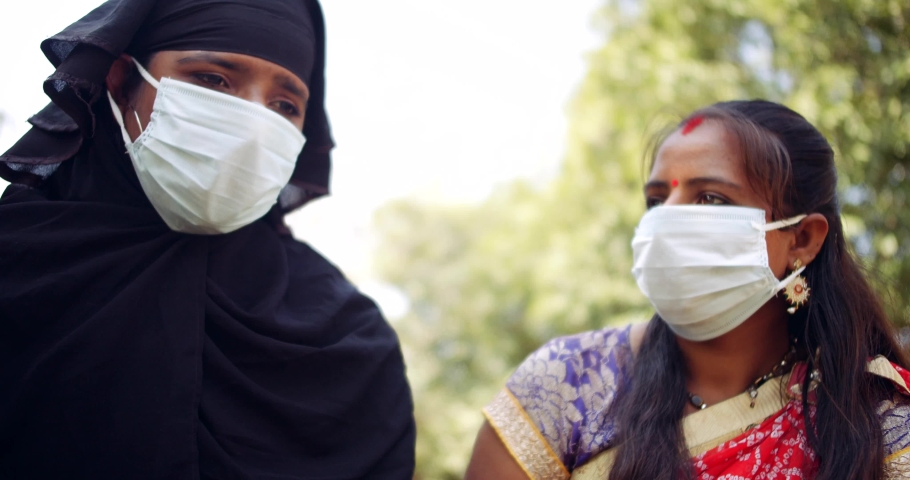 Slow-motion of two females friends with white surgical face mask outdoors public park garden nature, muslim woman in black burka nikab covered head and hindu lady in colorful red blue Sari
