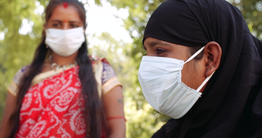 Slow-motion of two females friends with white surgical face mask outdoors public park garden nature, muslim woman in black burka nikab covered head and hindu lady in colorful red blue Sari