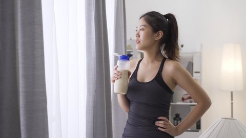 asian chinese woman in healthy lifestyle wearing sport clothes drinking bottle of milk at home by window after indoors workout. relax strong lady enjoy protein shake beverage in house living room.