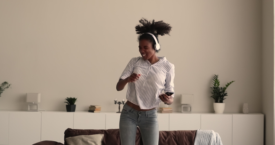 African 25s woman wear wireless headphones listen playlist of funky energetic favourite music, sing song jumping while dance get high on weekend at home alone. Hobby, free time use modern tech concept Royalty-Free Stock Footage #1065237148