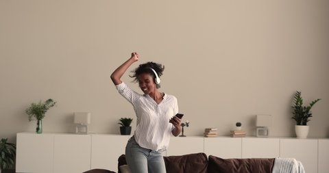 African 25s woman wear wireless headphones listen playlist of funky energetic favourite music, sing song jumping while dance get high on weekend at home alone. Hobby, free time use modern tech concept