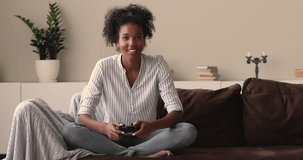 Young African ethnicity woman player sit on couch cross legged at home alone holding console game pad involved at exciting cool video game playing. Gaming, free time on weekend, funny activity concept