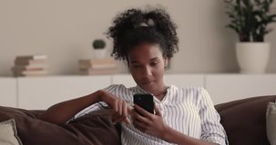 Modern technology every day usage, addiction, gadget overuse concept. African woman relaxing on couch at home spend free time using smartphone chatting online, browse web internet pages feels carefree