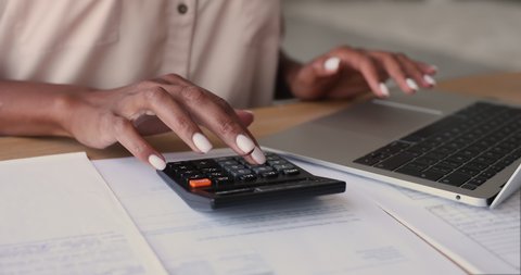 African woman sit at desk calculates costs using calculator and laptop, close up view. Female manage expenditures, control personal expenses, pay through e-bank app. Business accounting work concept