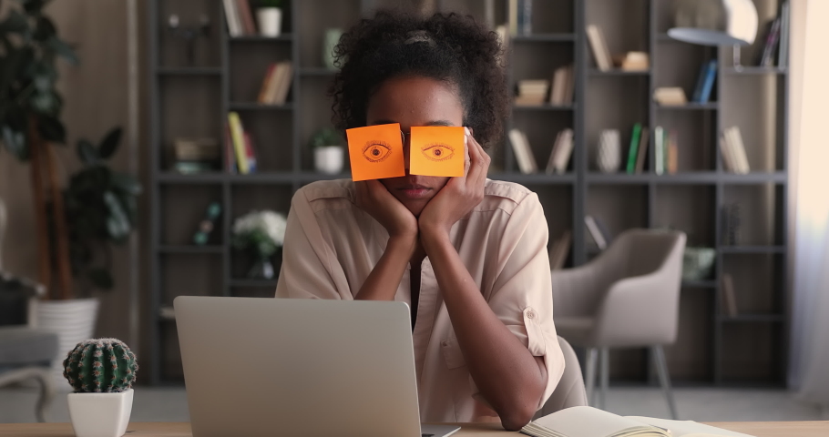Tired sleepy african business woman with orange adhesive notes on her eyes. Fatigued indifferent clerical female worker sit at desk feel bored due routine work. Overworked unmotivated employee concept | Shutterstock HD Video #1065238093
