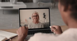 50s woman talk to colleague using video conference, laptop screen view over african female shoulder sit at desk take part in remote communication with elderly friend living abroad. Video call concept