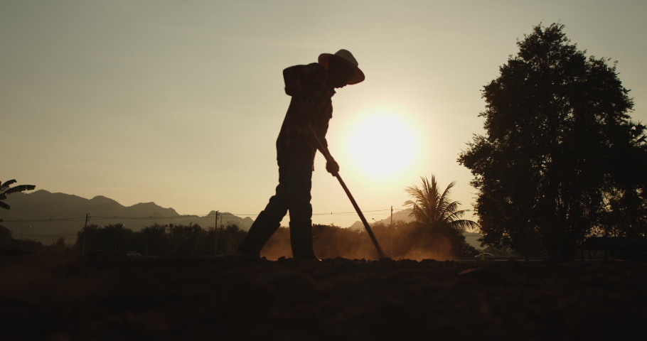 Slow motion silhouette scene of rural male Asian farmer digging the soil with a hoe to prepare for planting as the sun sets with sunlight passing through him beautifully. Royalty-Free Stock Footage #1065242803