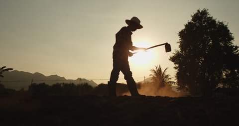 Slow motion silhouette scene of rural male Asian farmer digging the soil with a hoe to prepare for planting as the sun sets with sunlight passing through him beautifully.