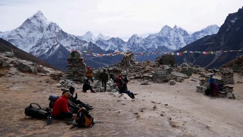 Trekkers resting at memorial for dead mountaineers with colorful prayer flags flying in wind near Lobuche, Khumbu, Nepal in Himalayas with majestic Ama Dablam. Text on mani stone: Buddhist mantra.