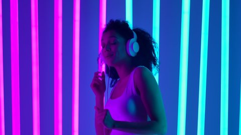 Portrait of a African American woman in big white headphones in a studio lit by bright blue and pink neon tubes. The girl is dancing enjoying the music from the headphones. Slow motion. Close up.