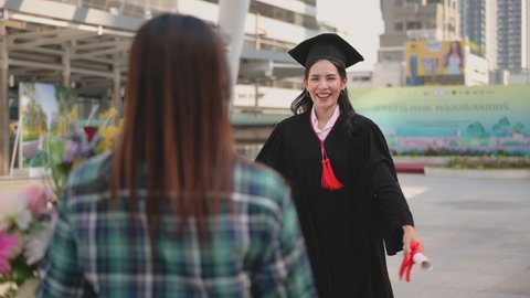 Happy beautiful young woman in graduation gowns with holding diploma are hugged mother by love and proud in city on outdoors. Asian mom embraces daughter with joy on graduation day and successful.