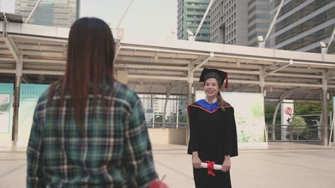Happy beautiful young woman in graduation gowns with holding diploma are hugged mother by love and proud in city on outdoors. Asian mom embraces daughter with joy on graduation day and successful.
