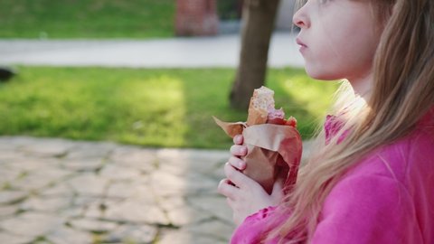 Side view of child in pink jacket who eats fast food.Cute little girl with long blonde hair eating hot dog on a outdoor bench.