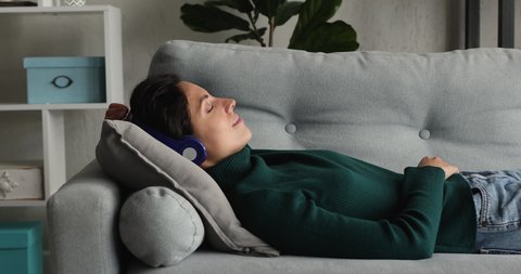 Tranquil woman lying on sofa cushion with eyes closed listen music through wireless headphones feels peaceful falling asleep. No stress, relaxation at home, quiet mind hearing audio meditation concept