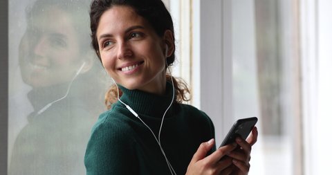 Close up woman relaxing seated on window sill hold smartphone listen favourite music through wired earphones smiles looks out window, rainy weather outside, comfort and warmth indoor, leisure concept