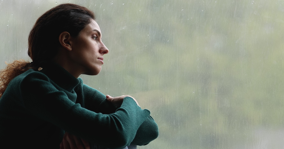 Sad woman sit on windowsill indoor feels upset looks outside through window rainy day gloomy weather, having melancholic mood, runs her finger on glass thinking about life troubles. Loneliness concept | Shutterstock HD Video #1065266062