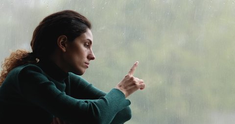 Sad woman sit on windowsill indoor feels upset looks outside through window rainy day gloomy weather, having melancholic mood, runs her finger on glass thinking about life troubles. Loneliness concept