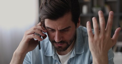Young man sit indoor talking on smartphone hearing bad news, office employee holding cellphone listen client complaints during phone conversation looking disappointed feel stressed, close up face view