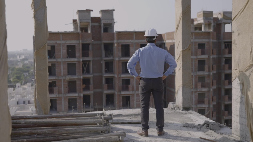 A movement shot of a young confident male asian civil engineer wearing hard hat and face mask standing on top of a under construction building turns around looks into the camera with crossed arms Royalty-Free Stock Footage #1065268189