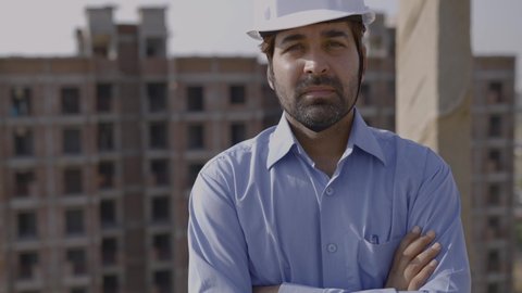 A movement shot of a young confident male asian civil engineer wearing hard hat and face mask standing on top of a under construction building turns around looks into the camera with crossed arms