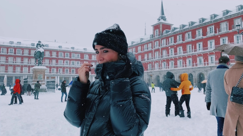 Pretty young caucasian girl looking at camera smiling during heavy snowfall in the city center.Plaza Mayor of Madrid (Spain) completely covered in snow.