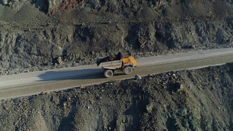 Drone flight together with yellow modern truck transporting mining ore. Heavy loaded vehicles are driving along dusty serpentine road up. Mining industry, abstract industrial landscape. Cement quarry