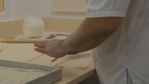Turkish man chef makes puff pastry for pkhlava. The cook cuts with a knife and rolls the dough with a wooden rolling pin on the table. Traditional oriental cuisine