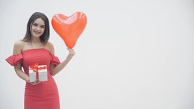Slow motion of joyful woman holding a gift box and heart balloon, smiling over a white background.