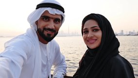 Couple spending time together in Dubai. Man and woman wearing kanura and hijab walking near the ocean and watching the skyline