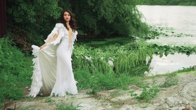 Fantasy happy woman queen in luxury long white dress stands posing by lake. Creative clothing cloak cape with natural feathers. Summer nature, water river green trees. Girl princess beauty river nymph